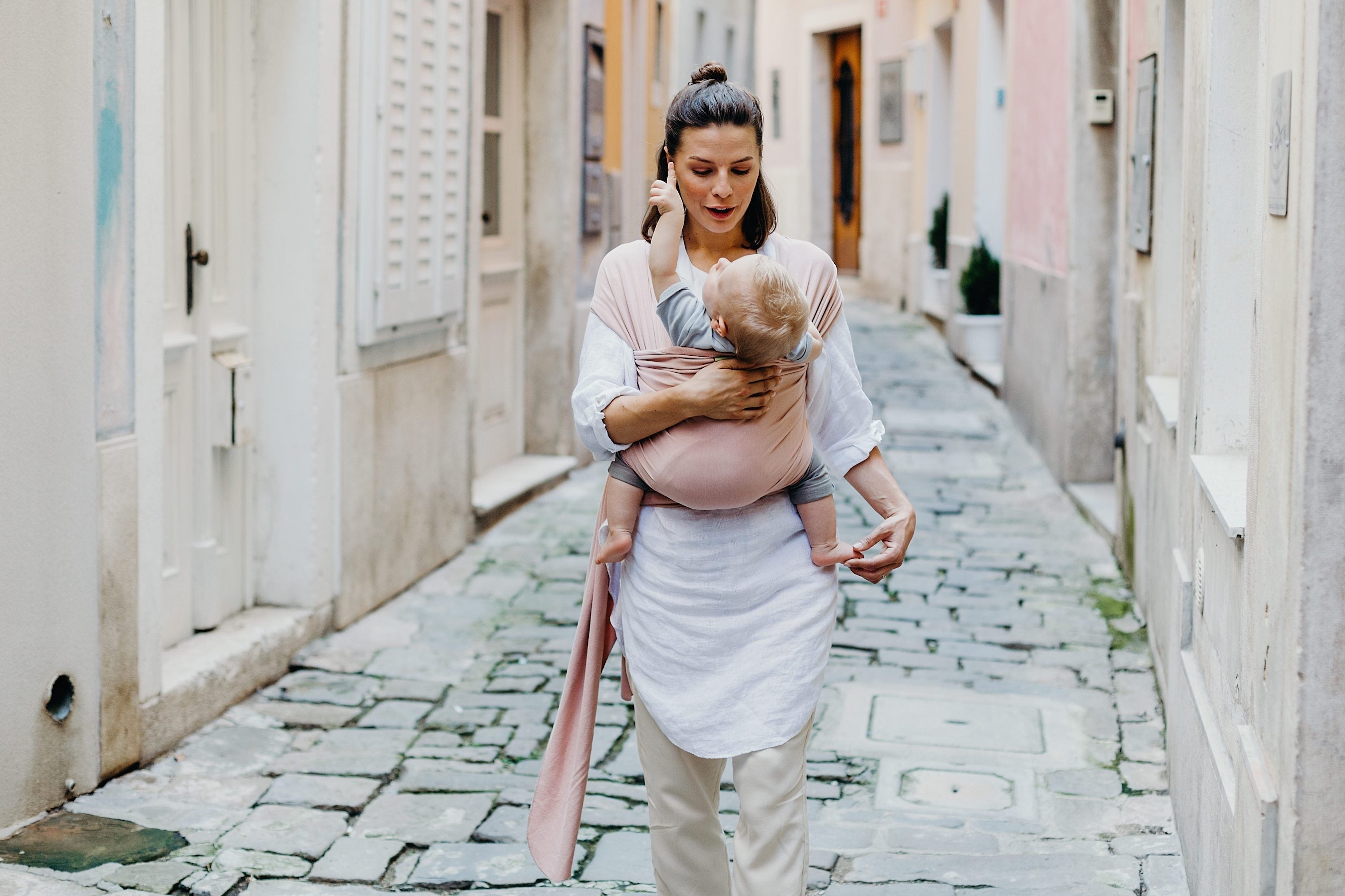 Mom making eye contact with her infant son while walking around a seaside town with her baby in a front carry position. Her son is pointing up in awe and she is wearing a summer outfit with a pale pink bamboo serenity wrap bloom.