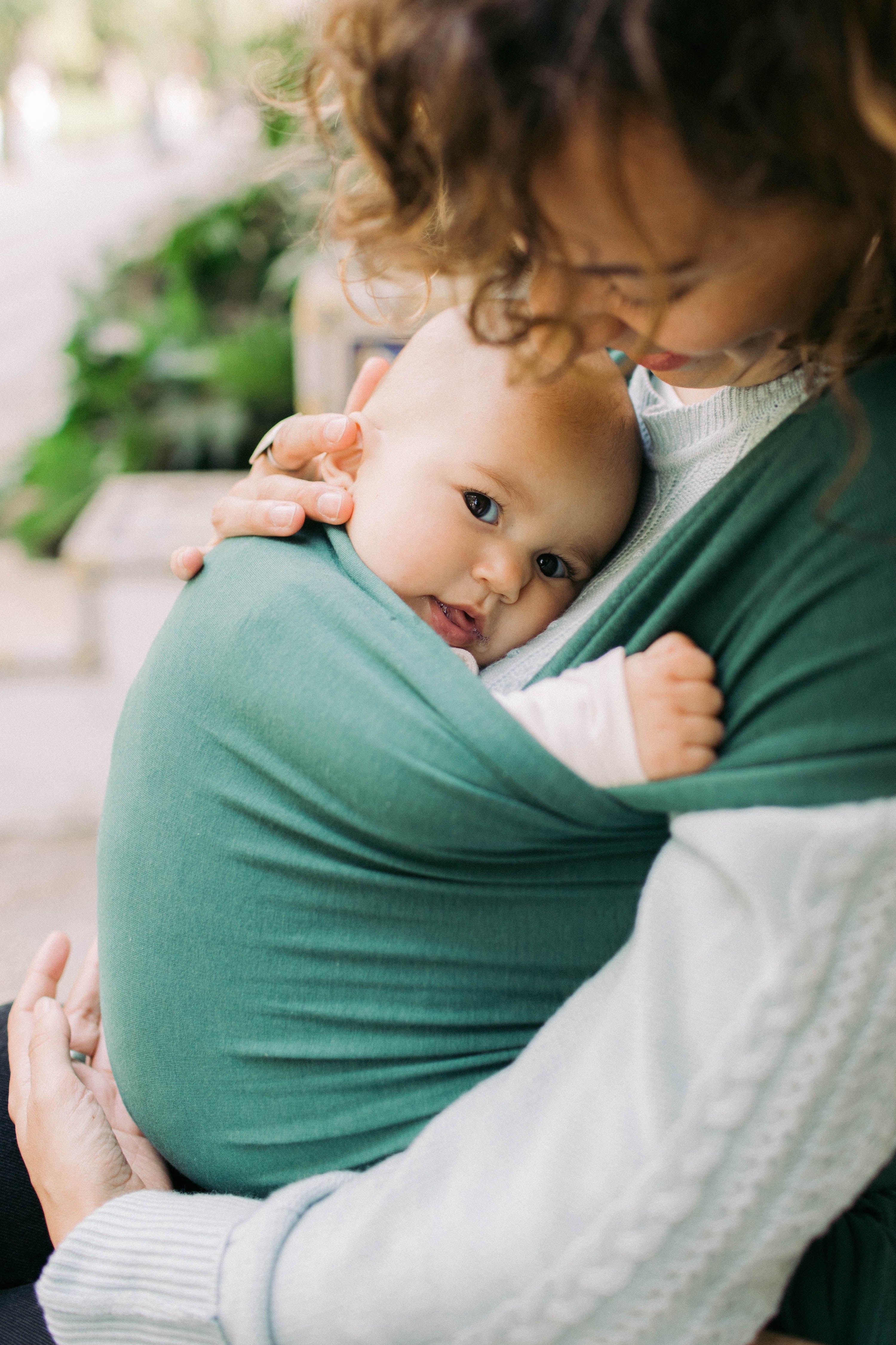 Brown curly haired mother is holding her baby daughter in the front carry in her rainforest serenity wrap.