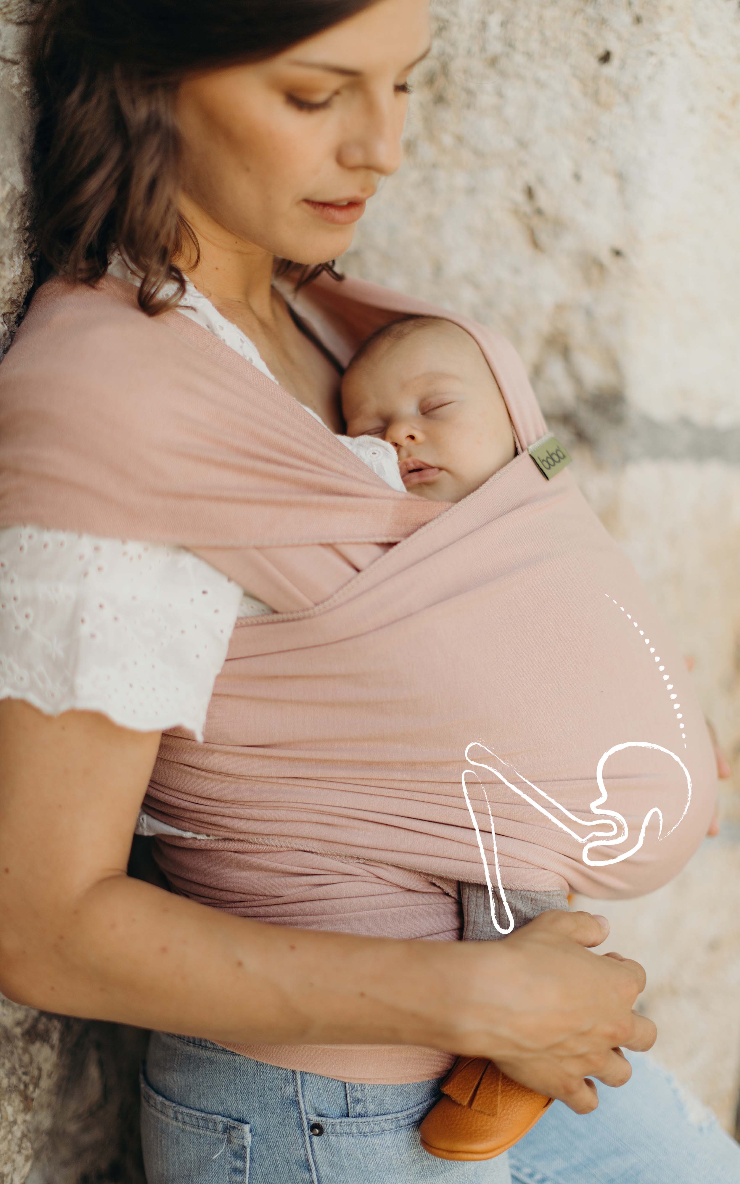 Mom wearing a white lace shirt and jeans has her sleeping baby girl in the bamboo serenity boba wrap. there is a line illustration of the baby's hips and spine in the natural ergonomic position over the photo.