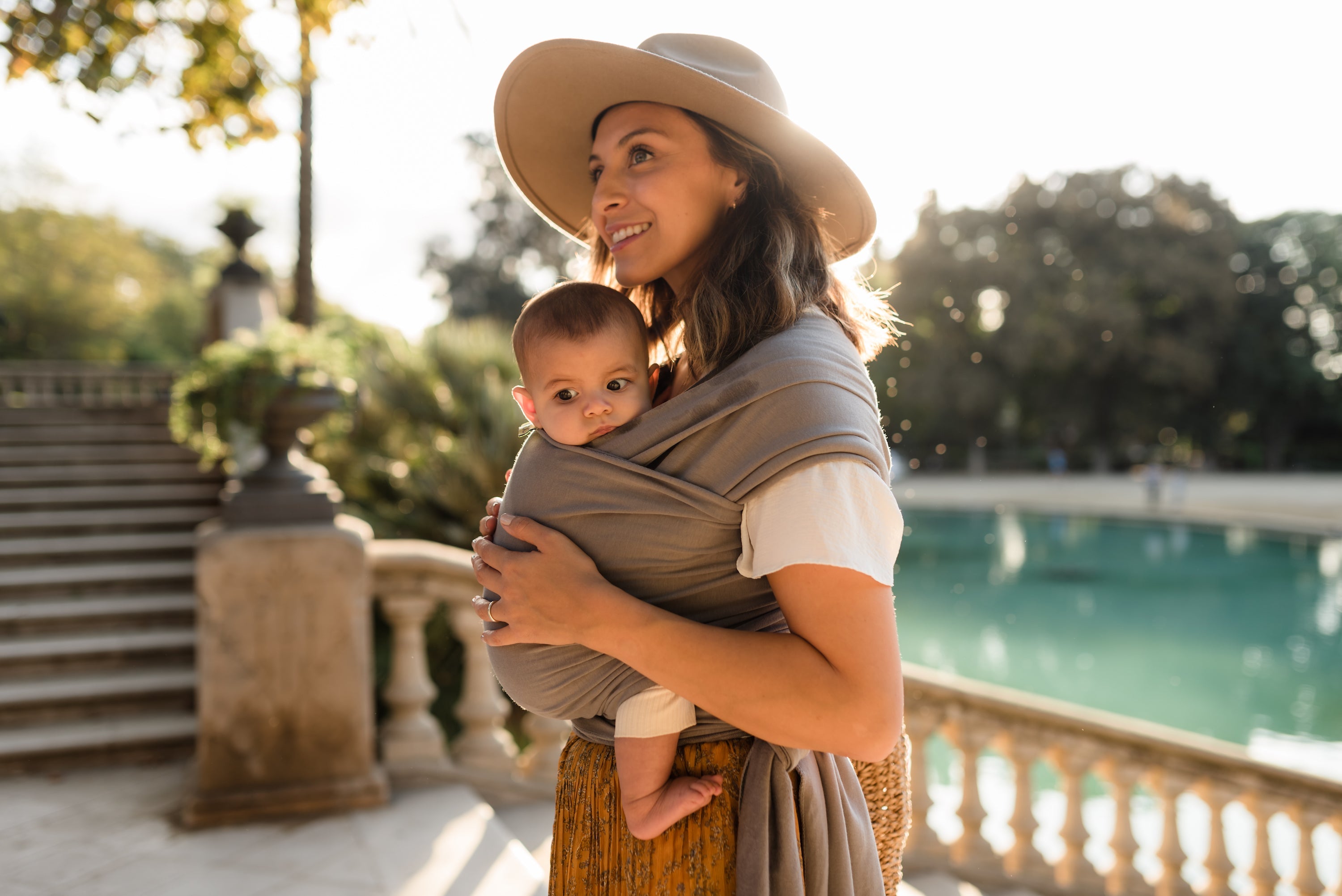 A happy mom is wearing her infant daughter in the gray boba wrap in an ergonomic front carry position. She is wearing a sun hat and standing outdoors in front of a fountain.