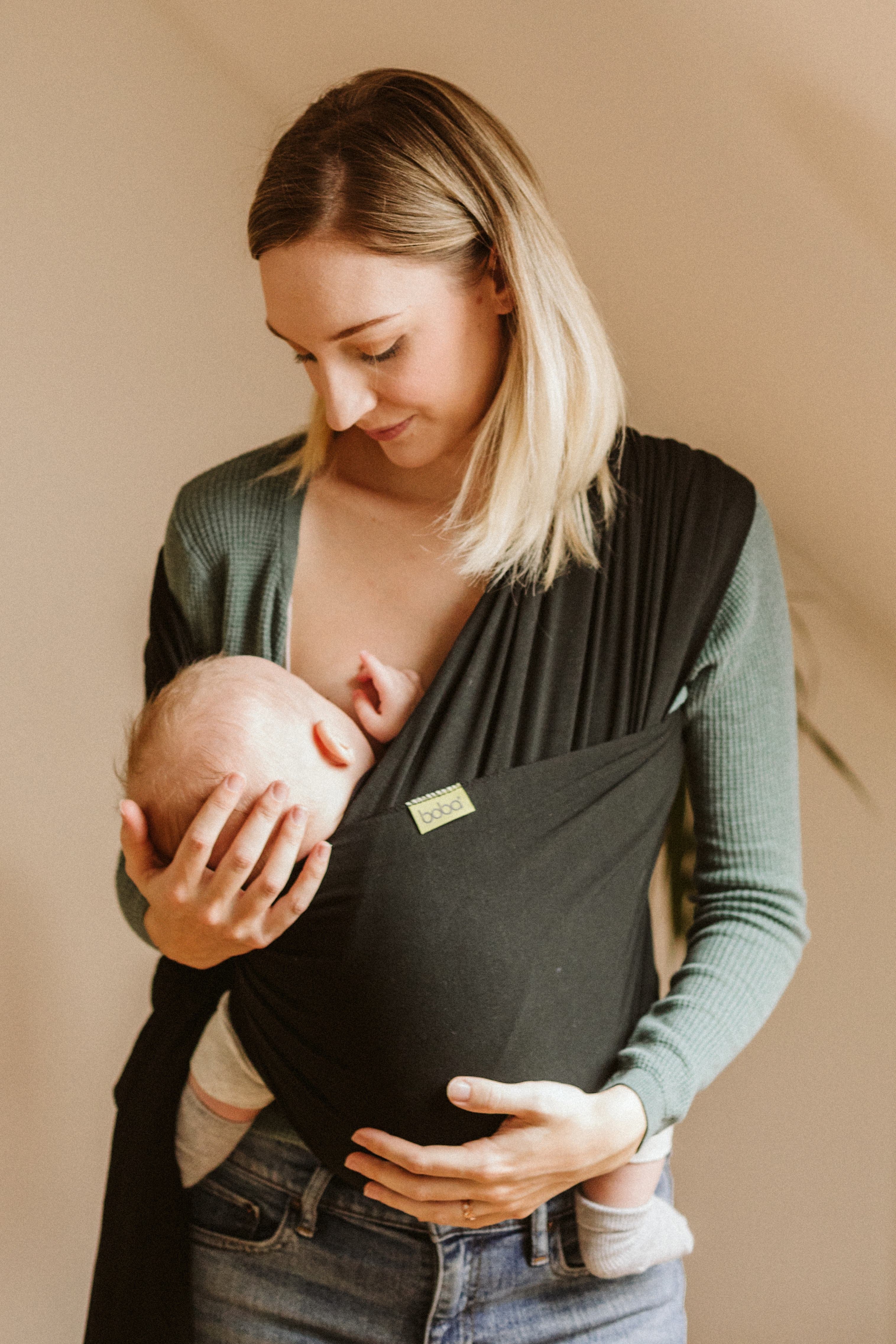 Calm and confident blonde young white mom is holding her baby boy's head while he nurses in the black serenity boba wrap made of bamboo.